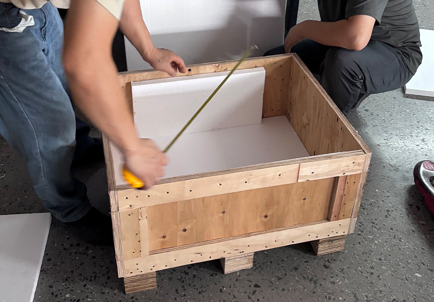 Workers measuring custom wooden crate with tape measure and foam padding for secure product packaging and shipping.