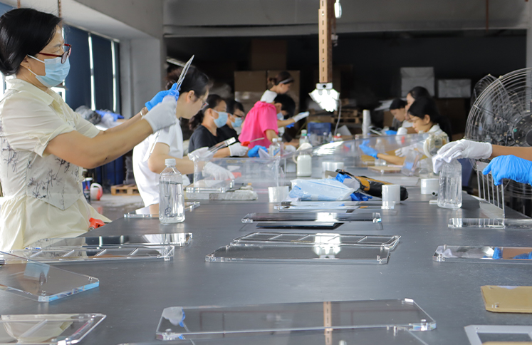 Factory workers wearing gloves and face masks cleaning, inspecting, and assembling custom clear acrylic sheets and organizers along a production workbench.