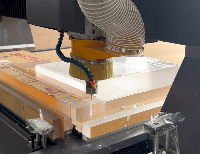 Industrial CNC router machine spindle milling and drilling thick solid acrylic blocks with dust collection brush in a factory workshop.